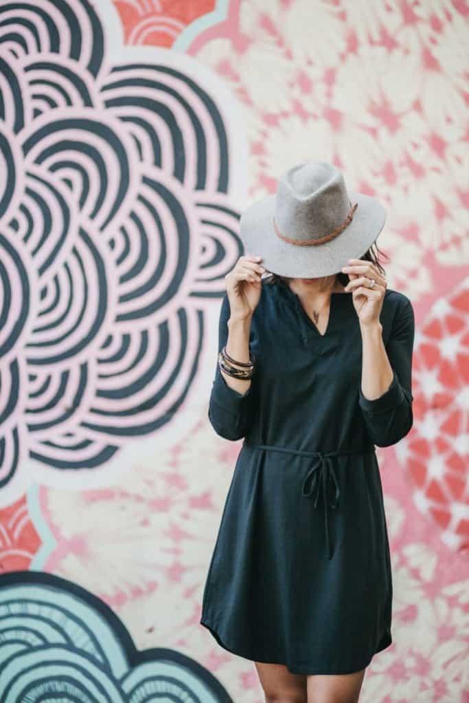 wearing a black dress with a gray fedora standing in front of city art 
