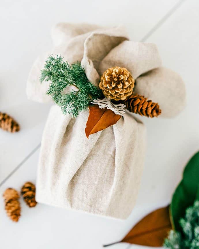 gift wrapped in white fabric with pine cones and greenery