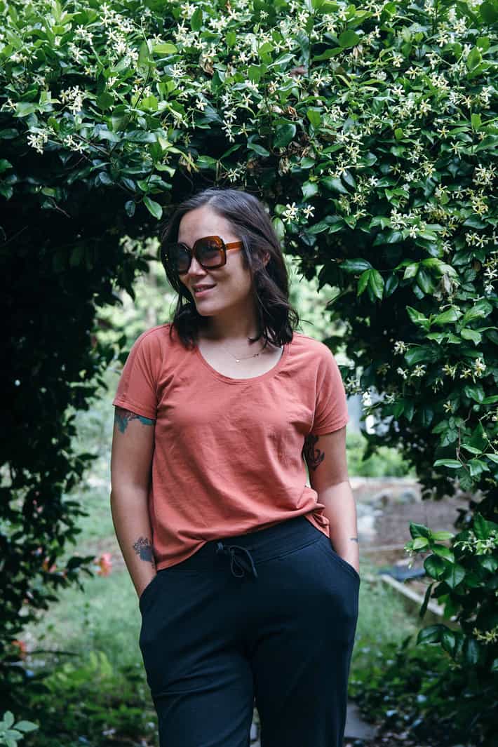Tanya wearing sunglasses and burnt orange Lightweight Lounge Tee with black joggers, standing in front of a flower garden.