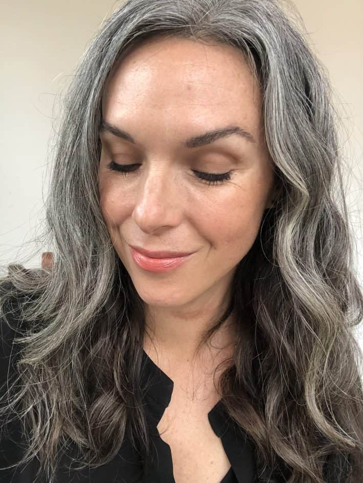 A woman  with wavy gray hair past her shoulders, smiles and looks down.