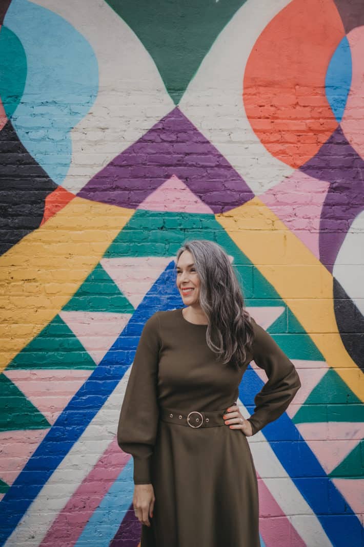 Lisa is standing in front of a multi-colored abstract painting on an outdoor wall, hand on hip.