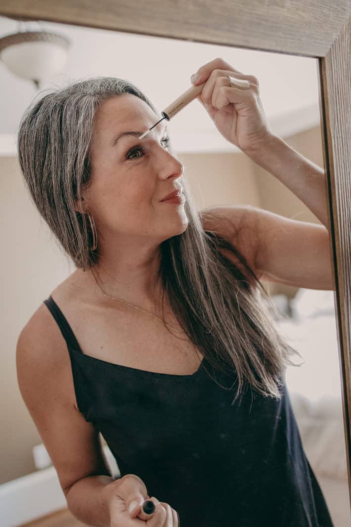 A woman stands in front of the mirror applying brow serum.