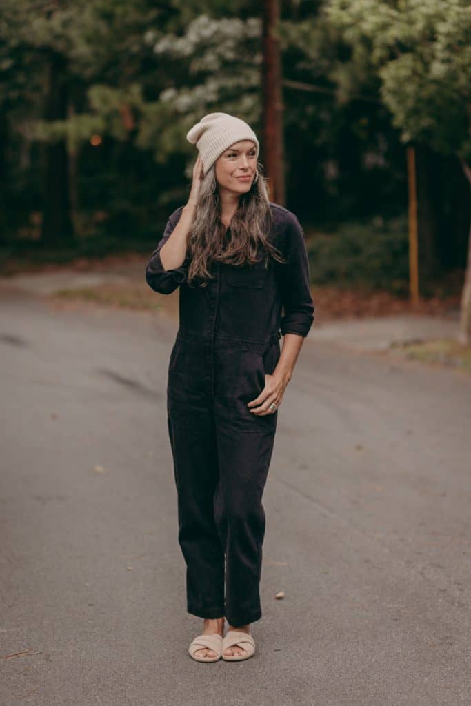 A woman stands in a black jumpsuit and knit hat.
