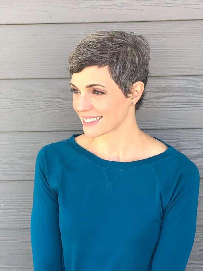 a woman in a blue top with a gray pixie cut stands in front of a gray sided wall