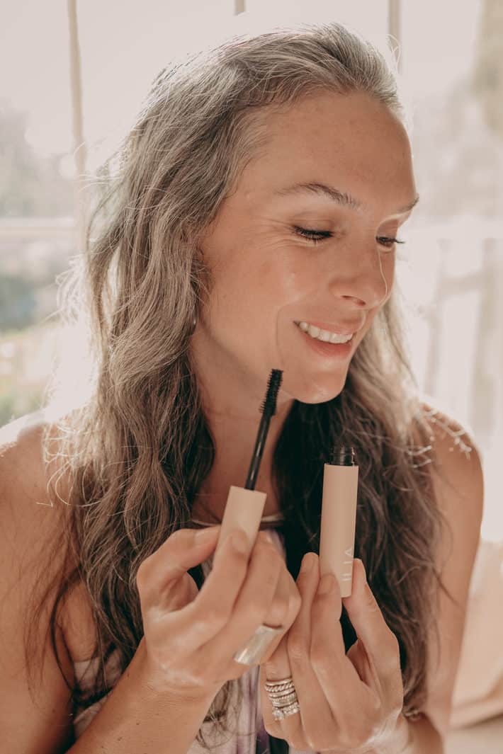 A woman holds opened tube of mascara in front of her face.