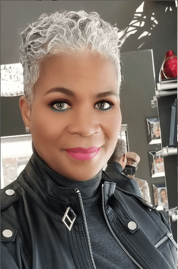 a woman with a gray pixie cut and pink lipstick is wearing a leather jacket at the hairstylist