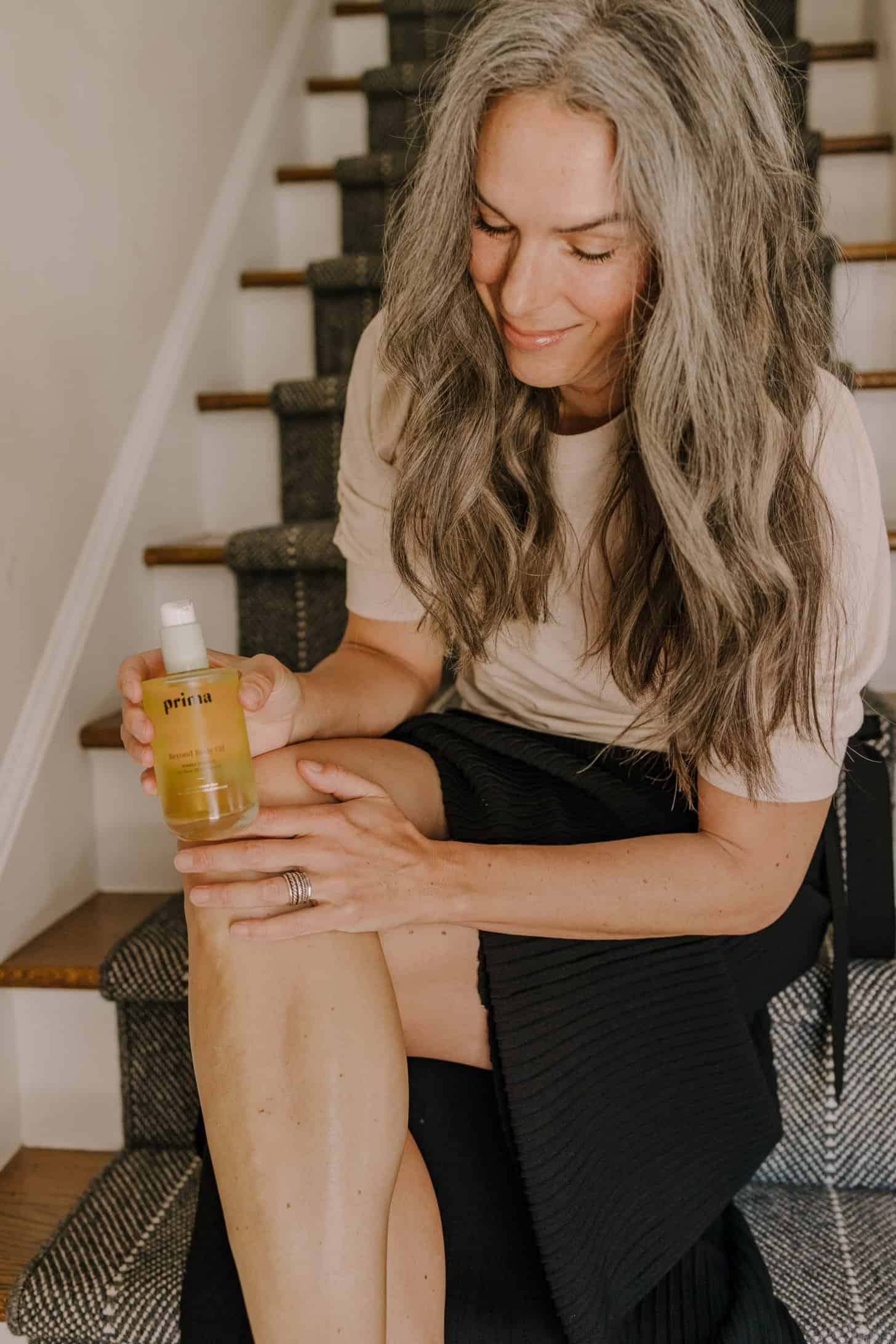 a woman applies prima body oil to her leg while sitting on the stairs