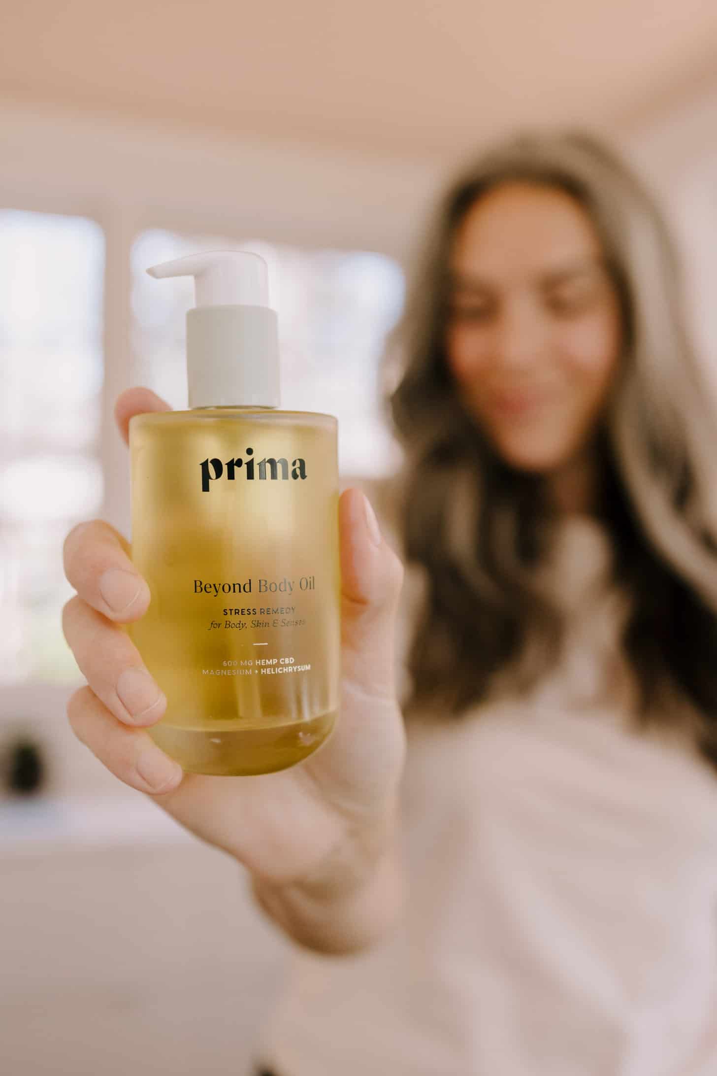a bottle of prima beyond body oil is held up to the camera while a woman stands in the background