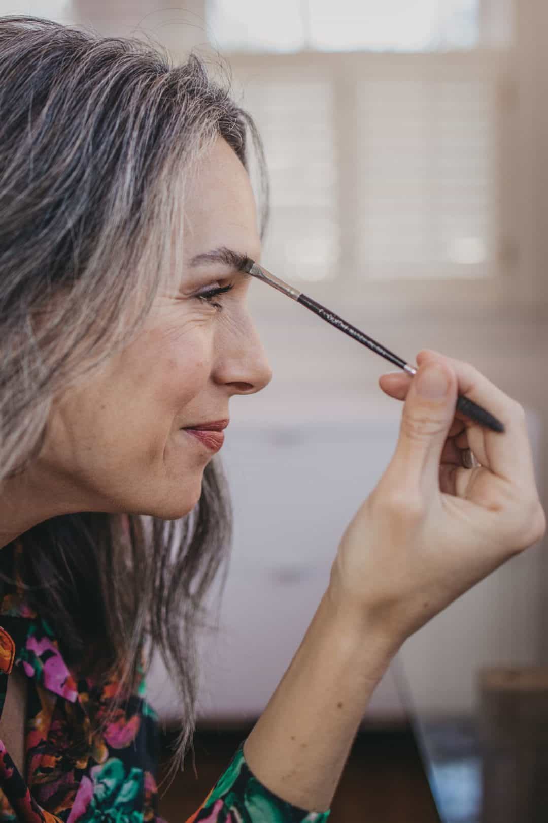 a sideshot of a woman applying makeup to her eyebrow while smiling