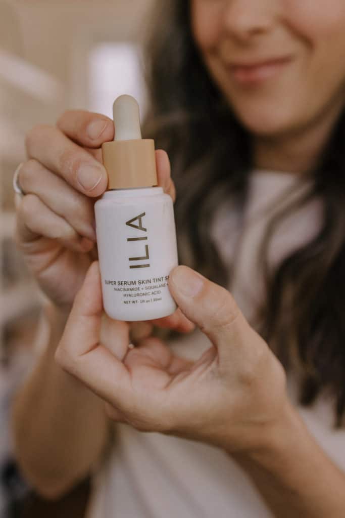 a woman stands in the background while holding up a bottle of ILIA super serum skin tint