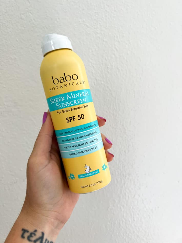 a hand holds up Babo Botanicals Sheer Mineral Sunscreen Spray SPF 50.