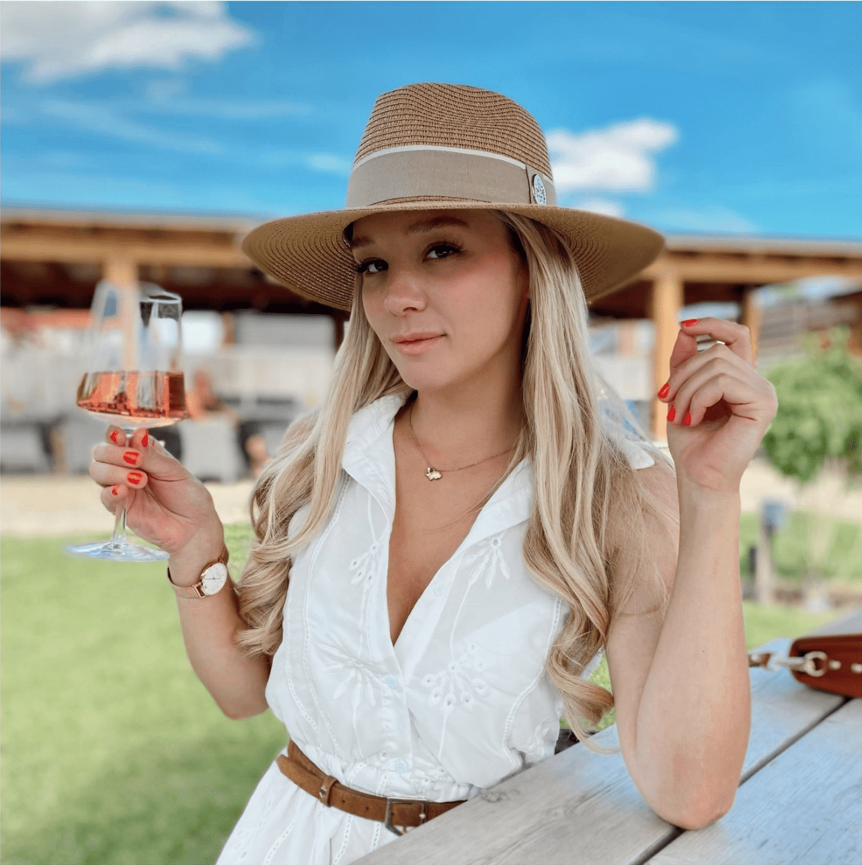 a woman with long blonde hair holds a glass of rose and wears a hat