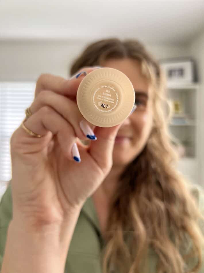 A woman with long curly hair holds up the bottom of ILIA True Skin Serum Foundation to the camera