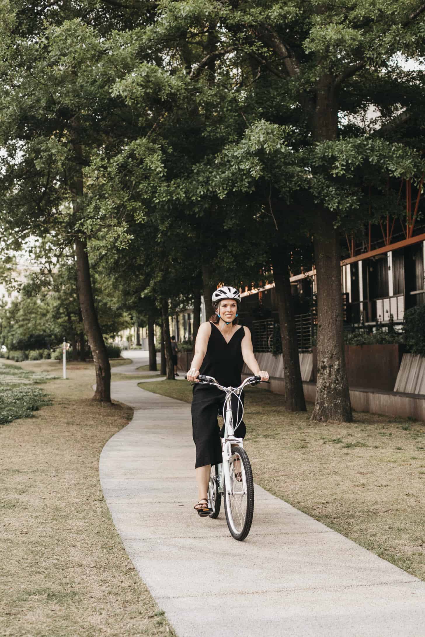 a woman in a black jumpsuit rides a bike down a tree lined path