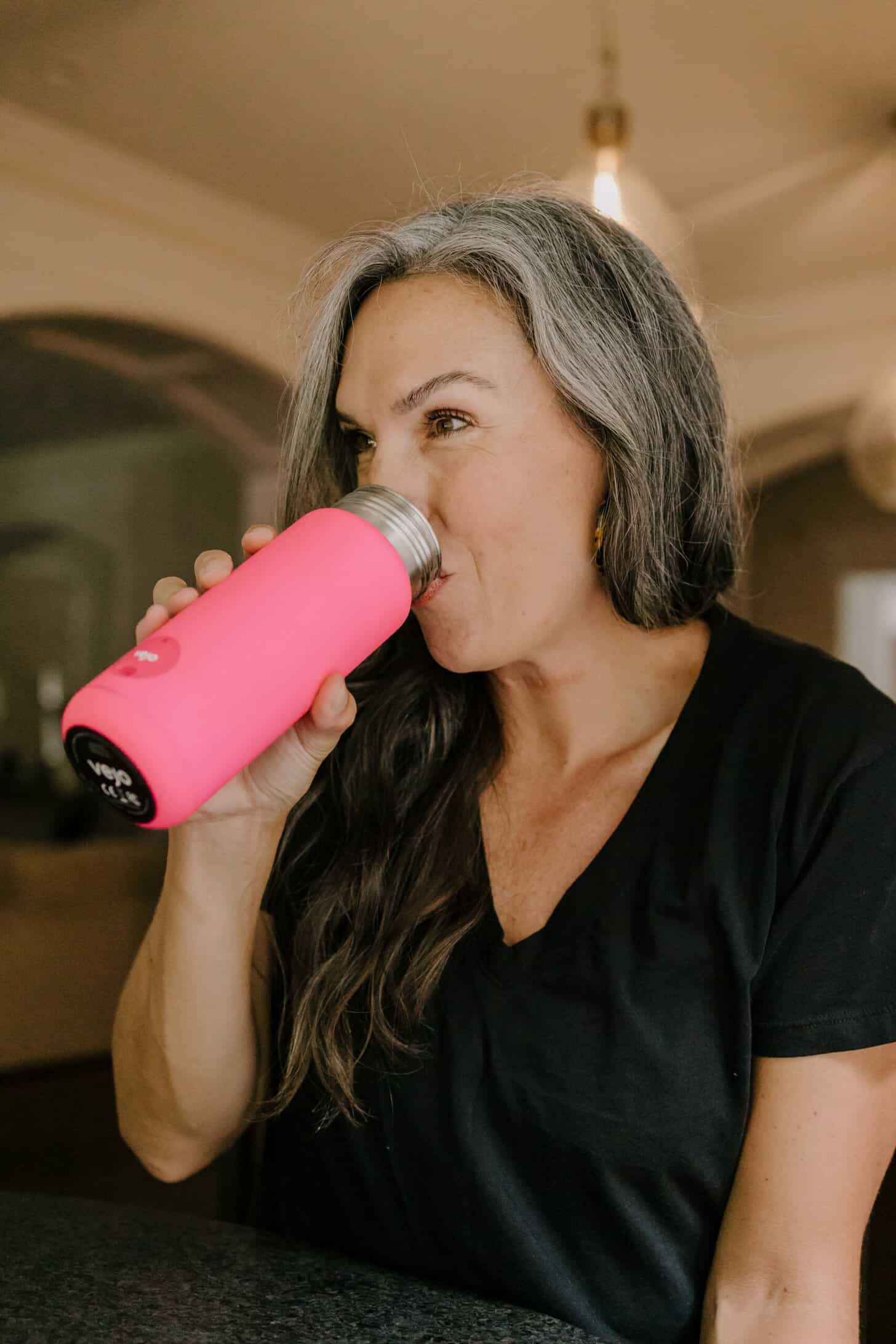 a woman with long wavy gray hair drinks from a hot pink portable blender