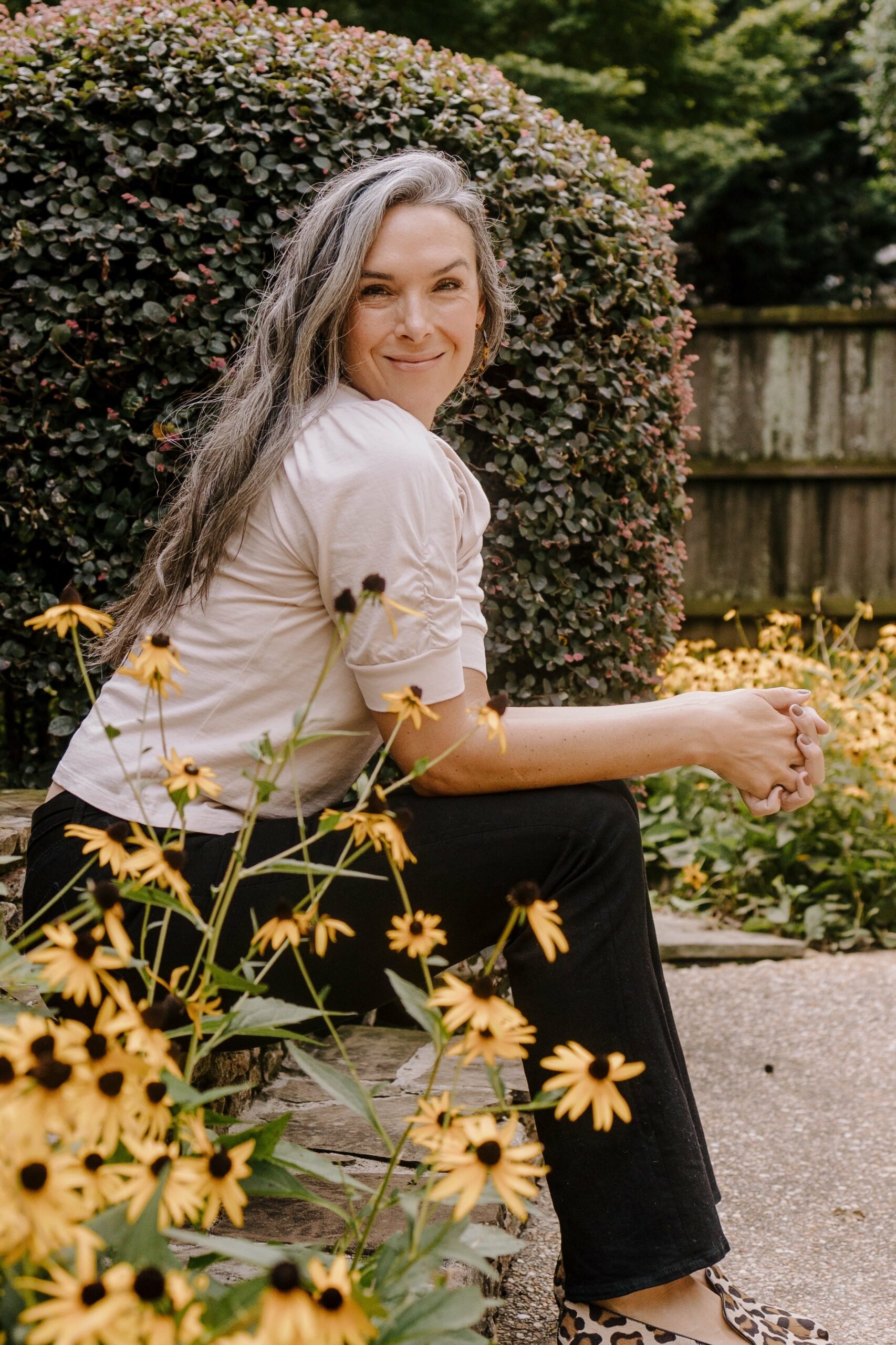 a woman with long wavy hair is seated near black eyed susan flowers in bloom