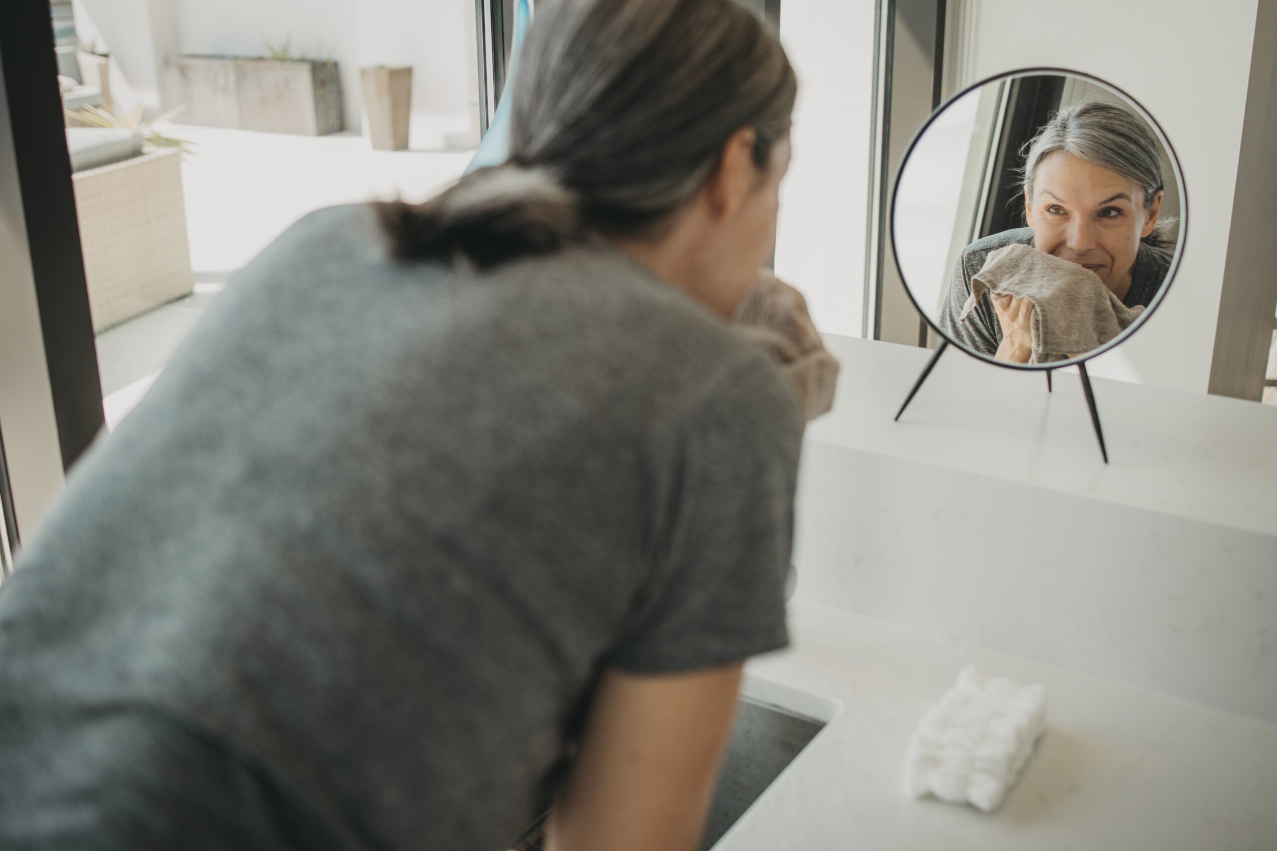 a woman drys her face with a towel while looking into a tabletop mirror