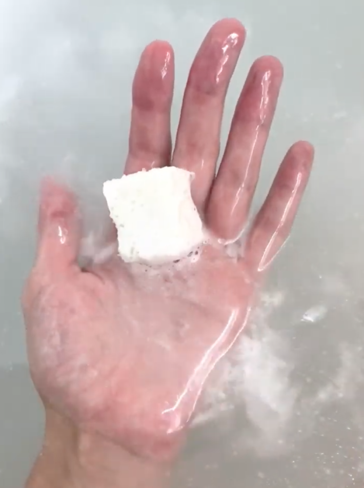a hand holds up a fizzing bath gem from Prima above a tub of water