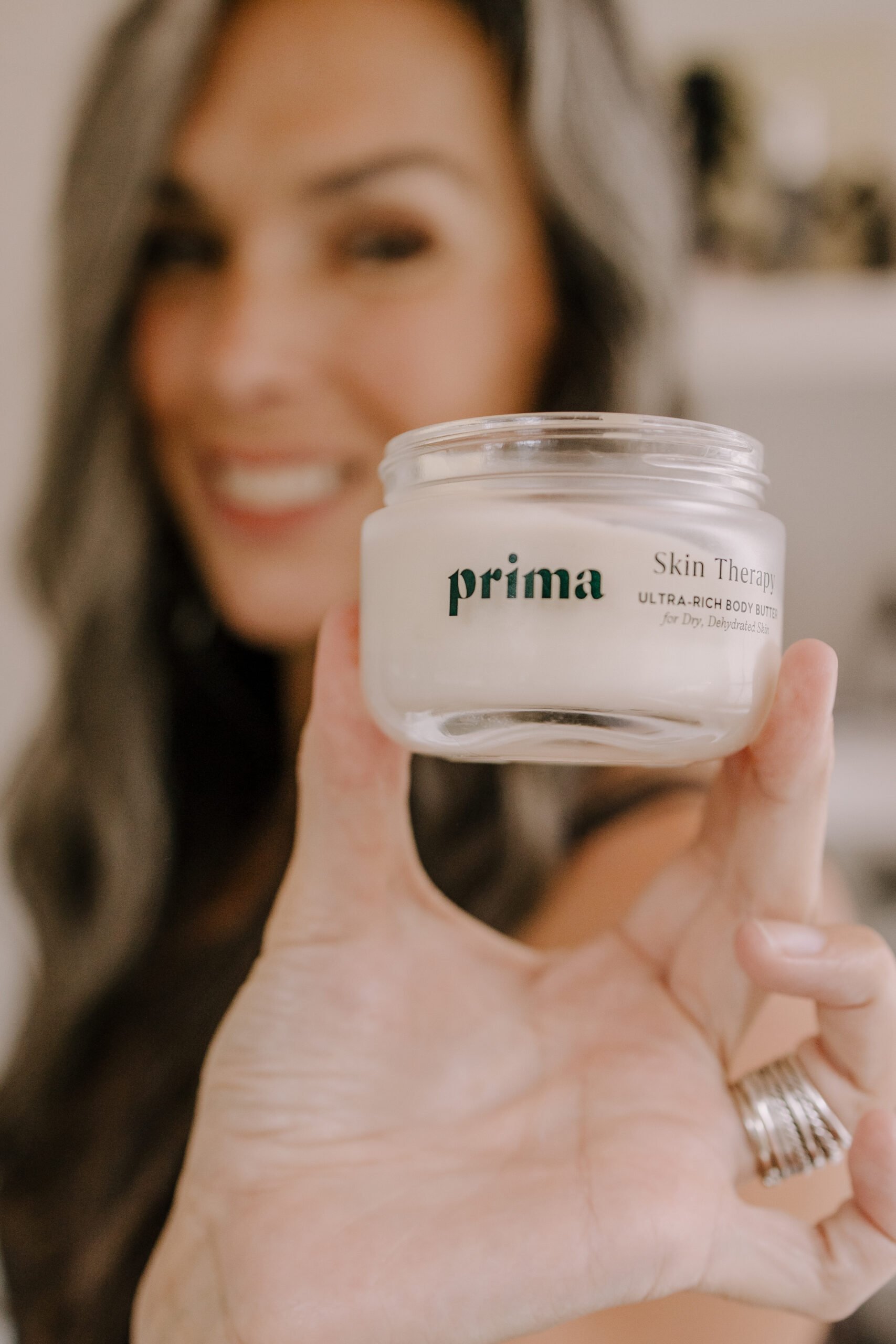a woman is blurry in the background while in the foreground is an opened tub of Prima's Skin Therapy that the woman is holding up