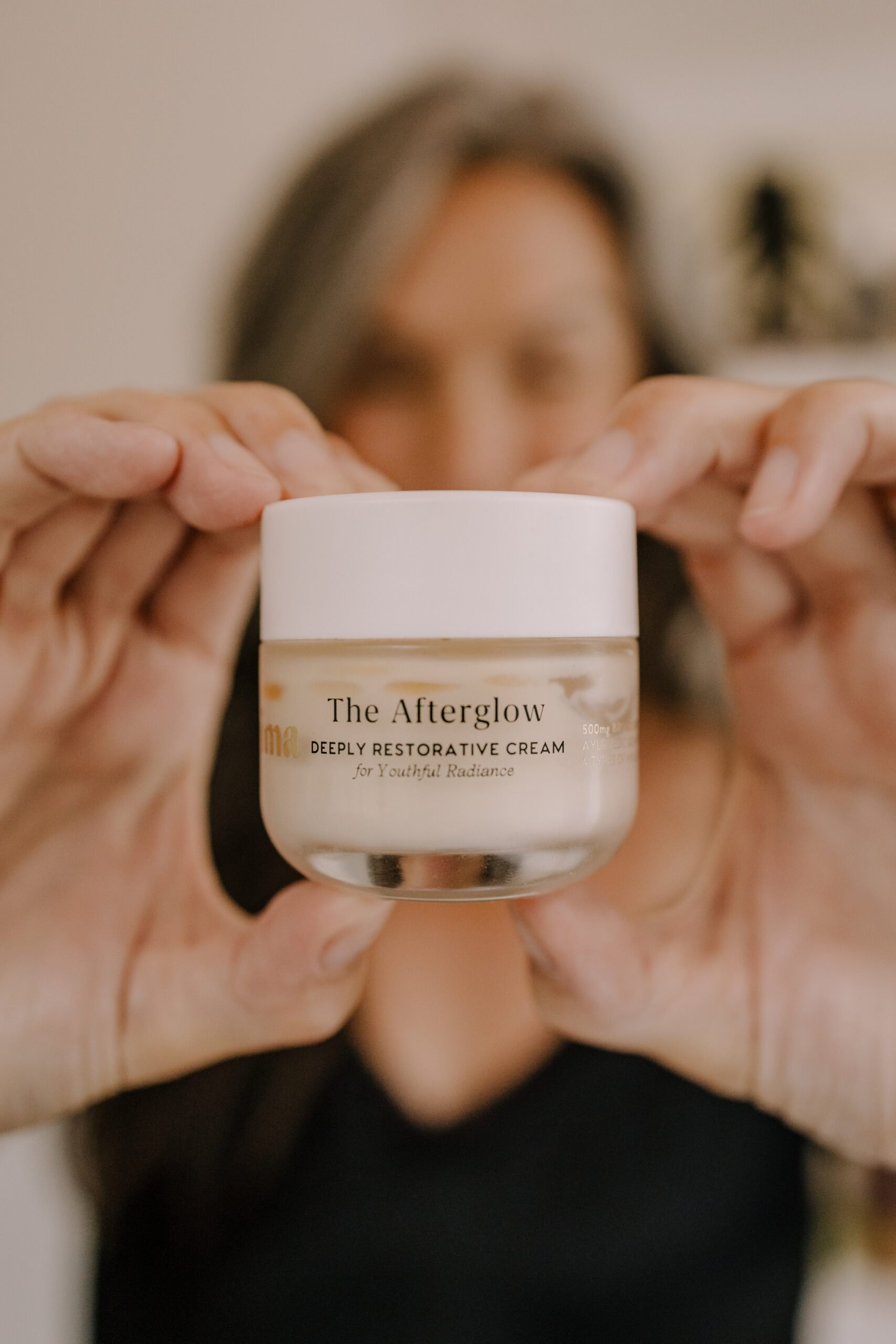 a woman in the background holds up a tub of Prima's The Afterglow moisturizer