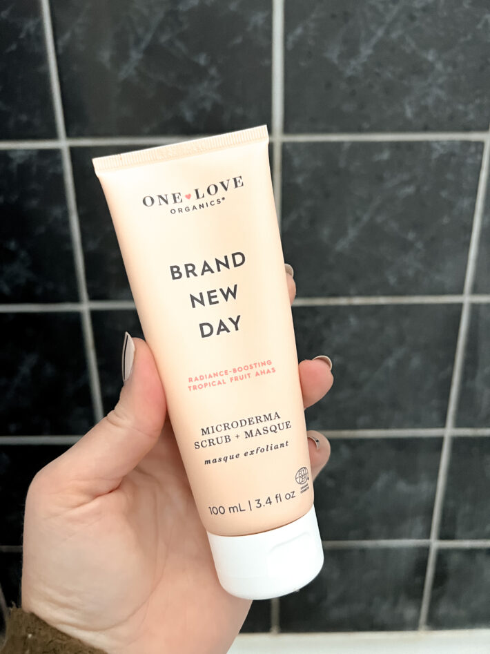 A hand holds up a tube of Brand New Day Face scrub by One Love Organics.