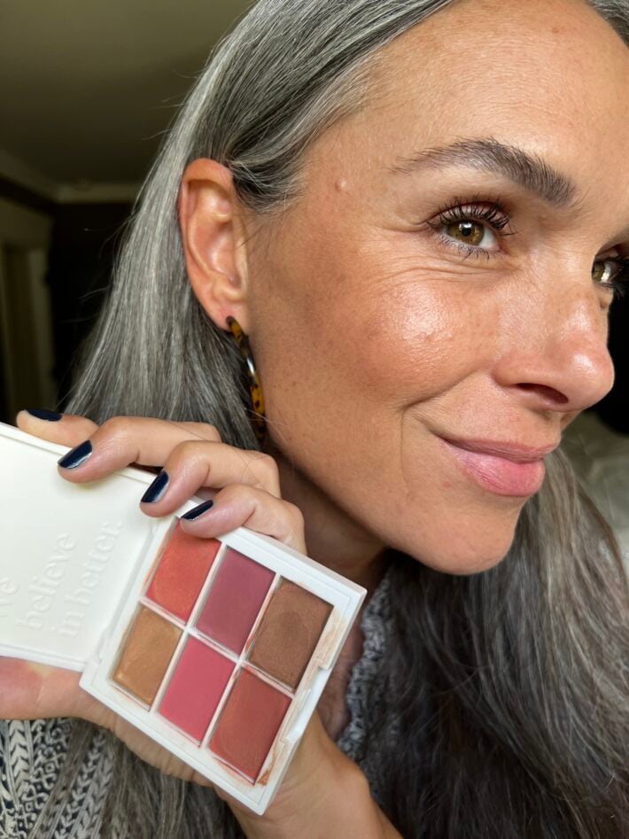 A woman holds up the ILIA multi-palette while showing it applied on her face as well.