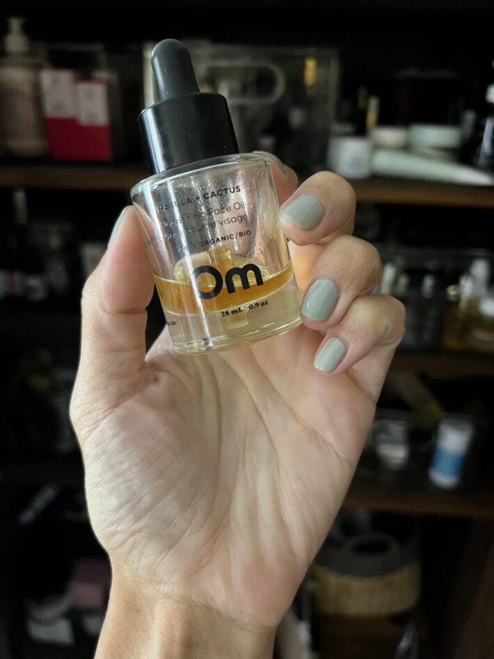 A hand with painted nails holds up a bottle of Om facial oil.