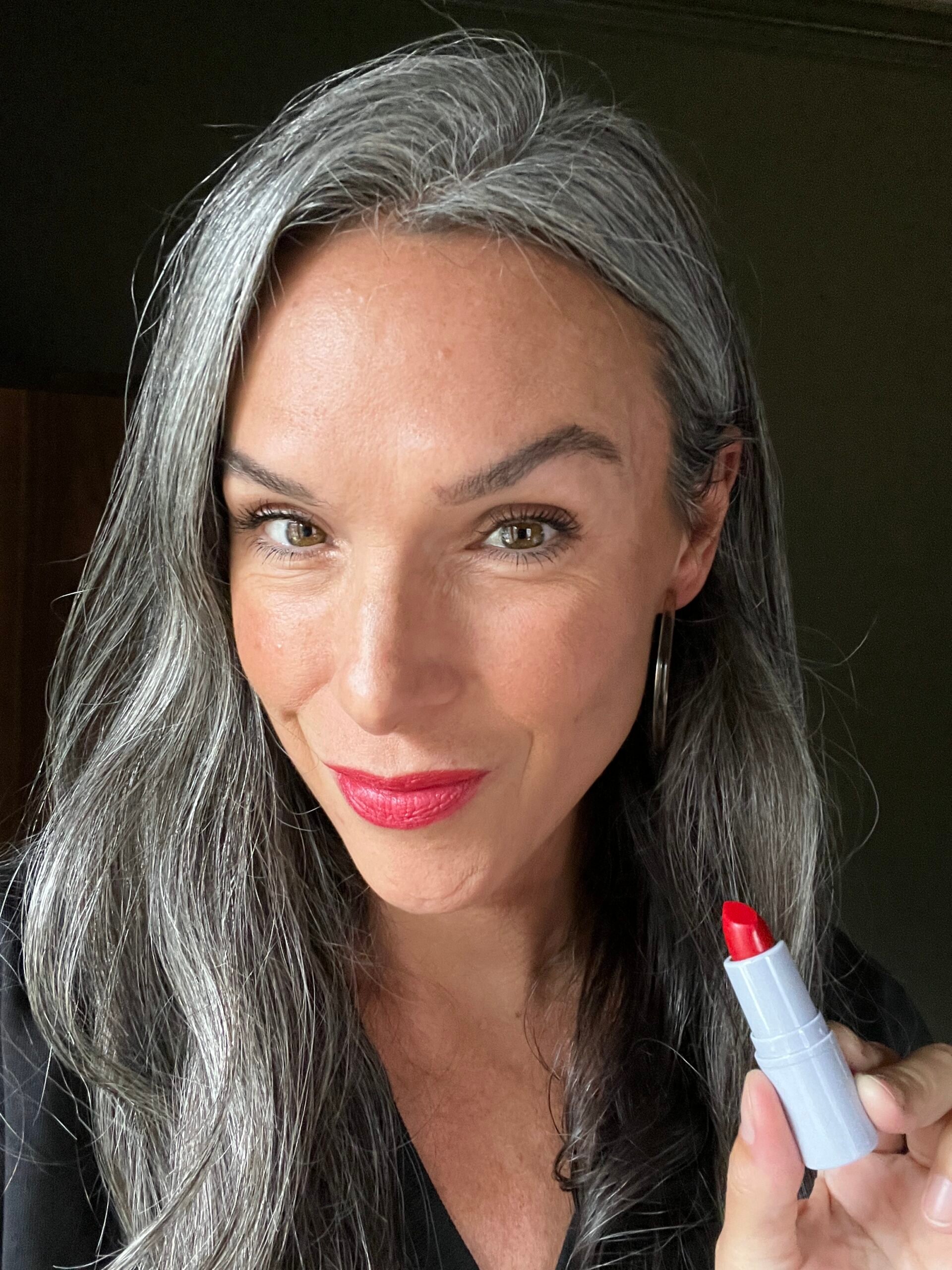 A woman wears Clover Comfy Matte Lipstick in Retro on her lips and holds up a tube of the lipstick.