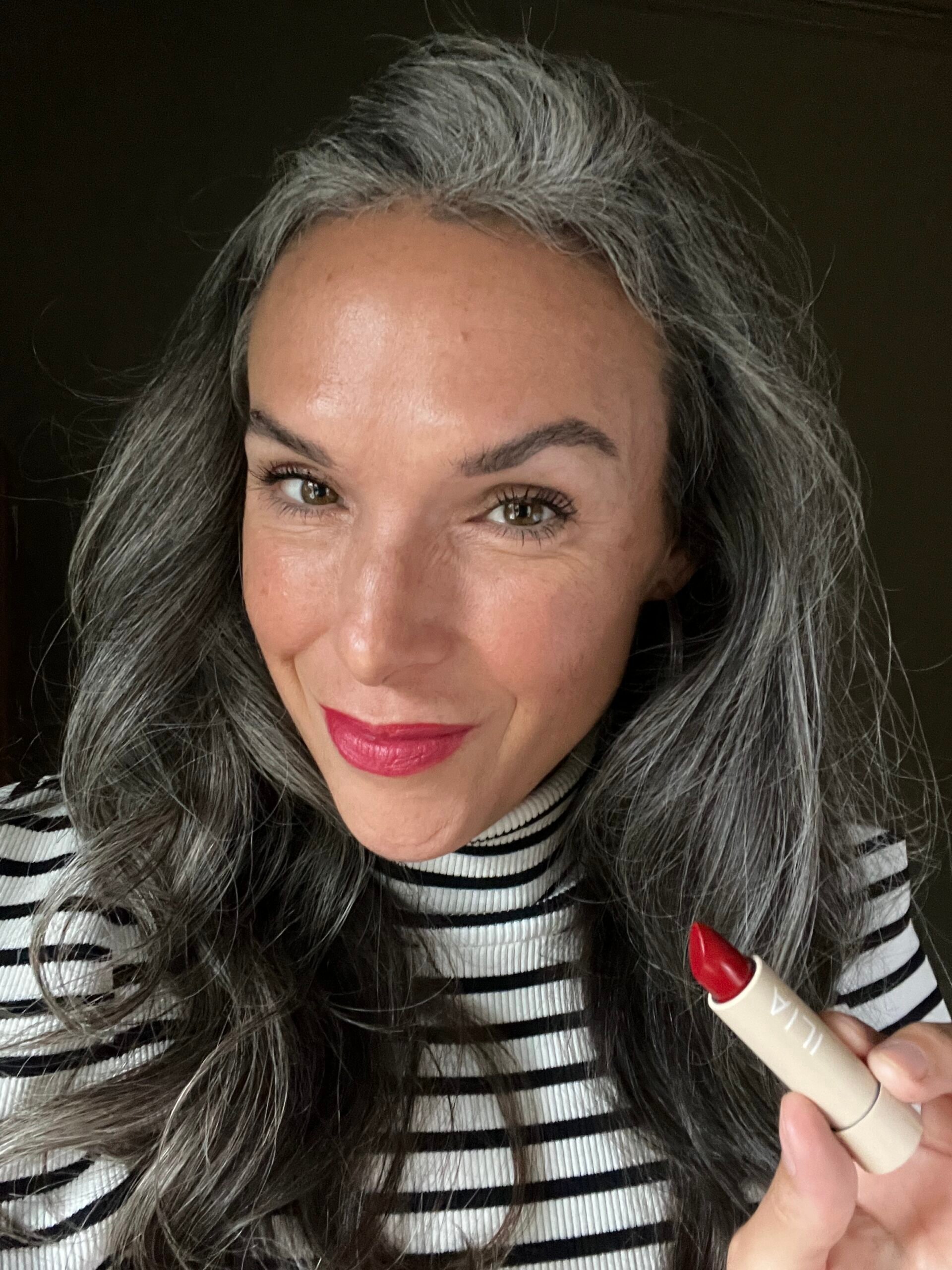 A woman with long gray hair holding ILIA lipstick.