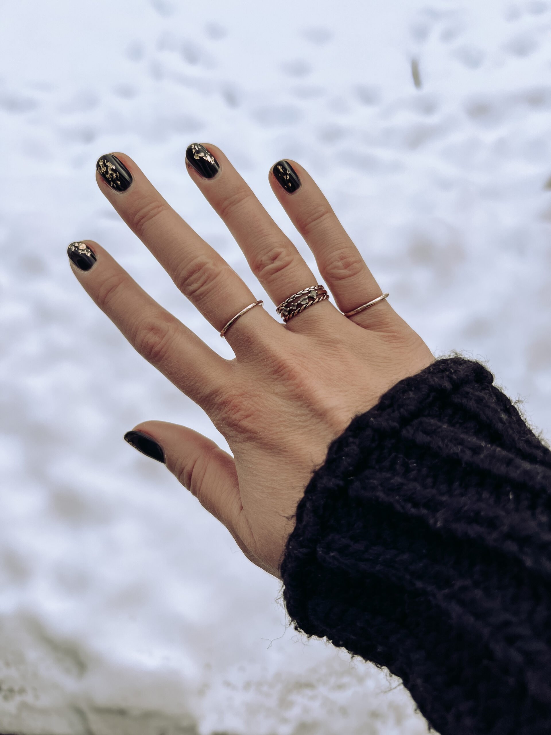Black painted nails on hand are held in front of a snowy background. 