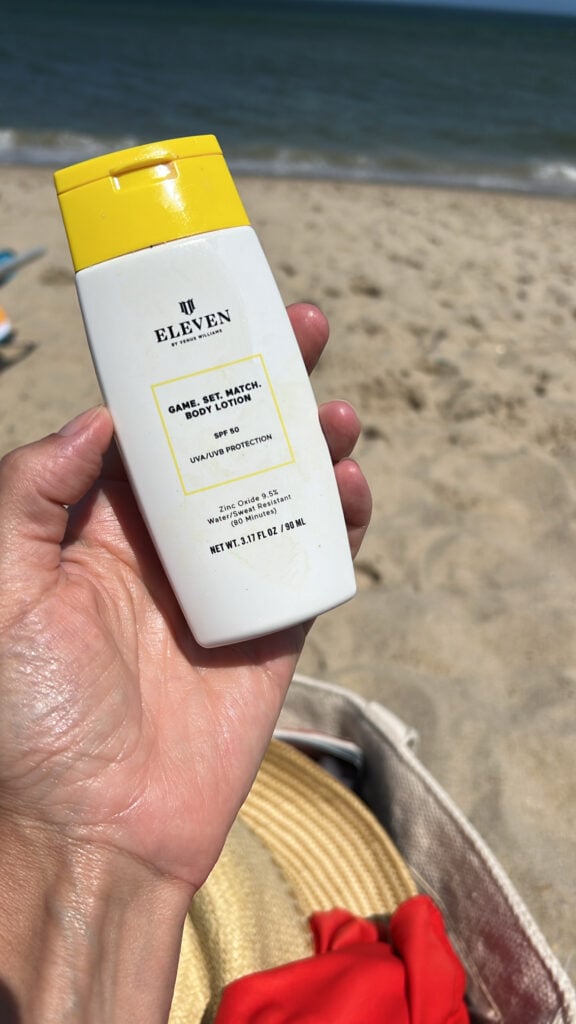 A bottle of Eleven Sunscreen held up on a beach.