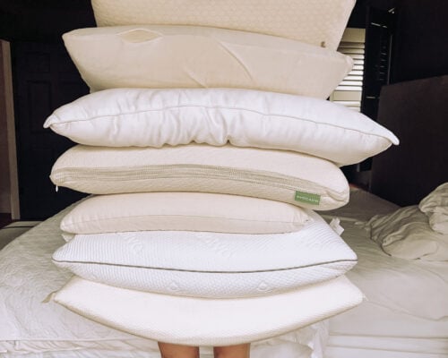 A stack of pillows is held up in front a person with only their legs visible.