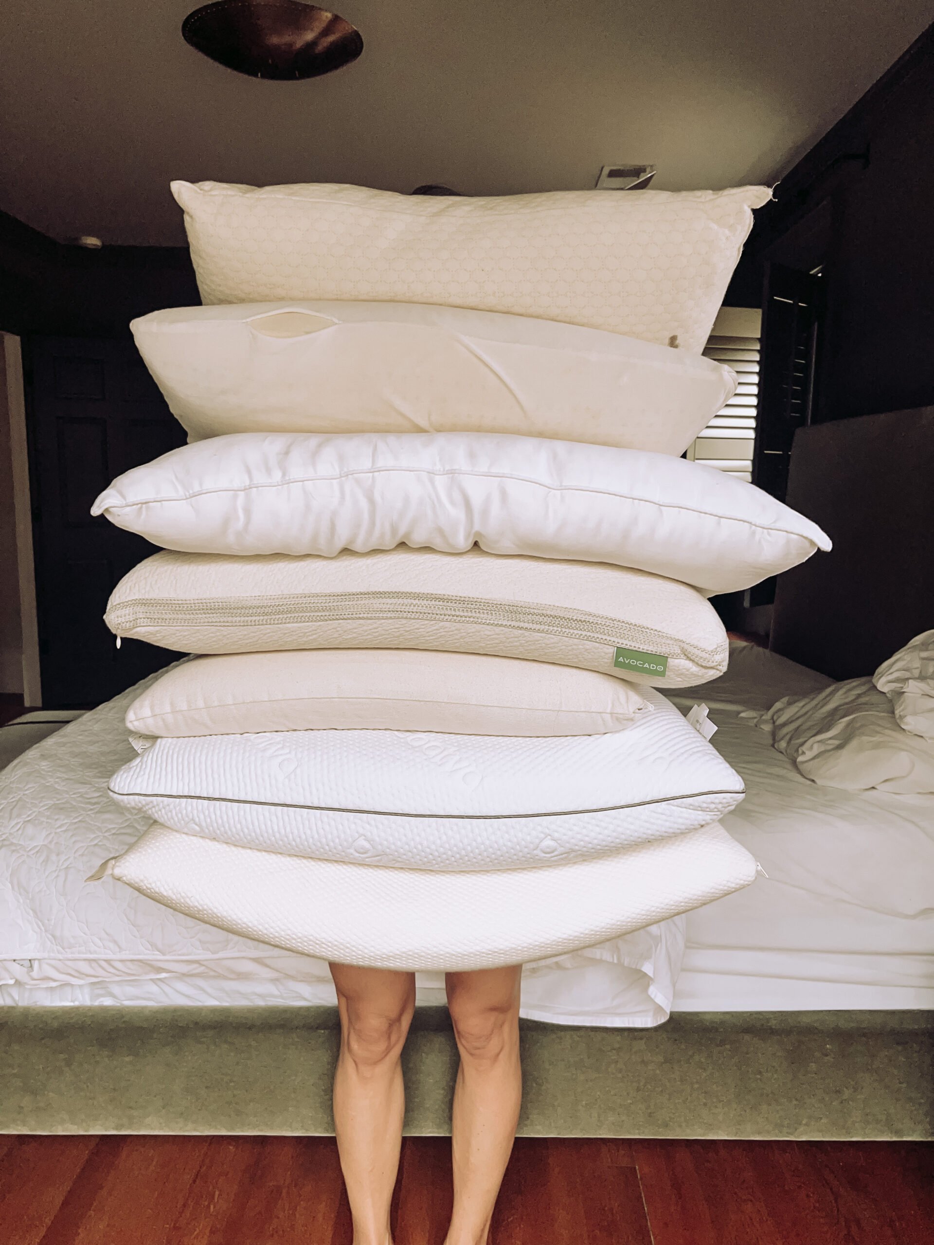 A stack of pillows is held up in front of a person so that only their legs are visible.