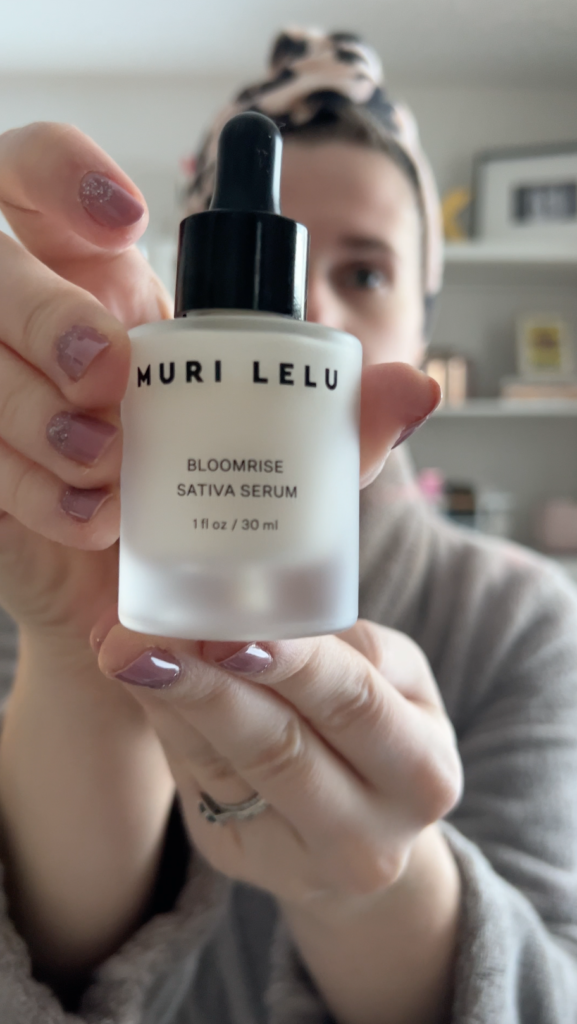 A woman holds up a bottle of Muri Lelu Bloomrise Sativa Serum.