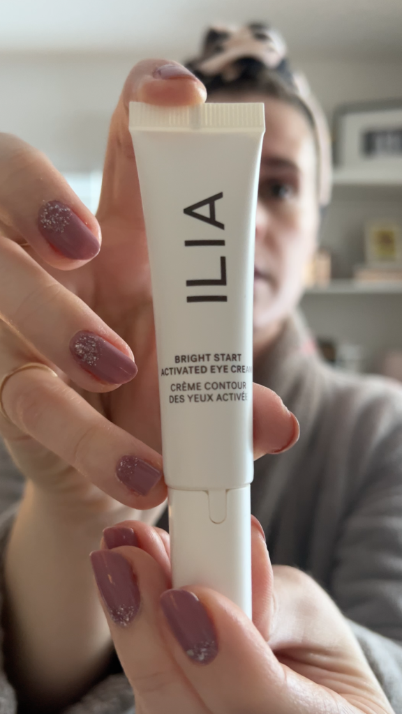 A woman holds up a tube of ILIA Bright Start Eye Cream.