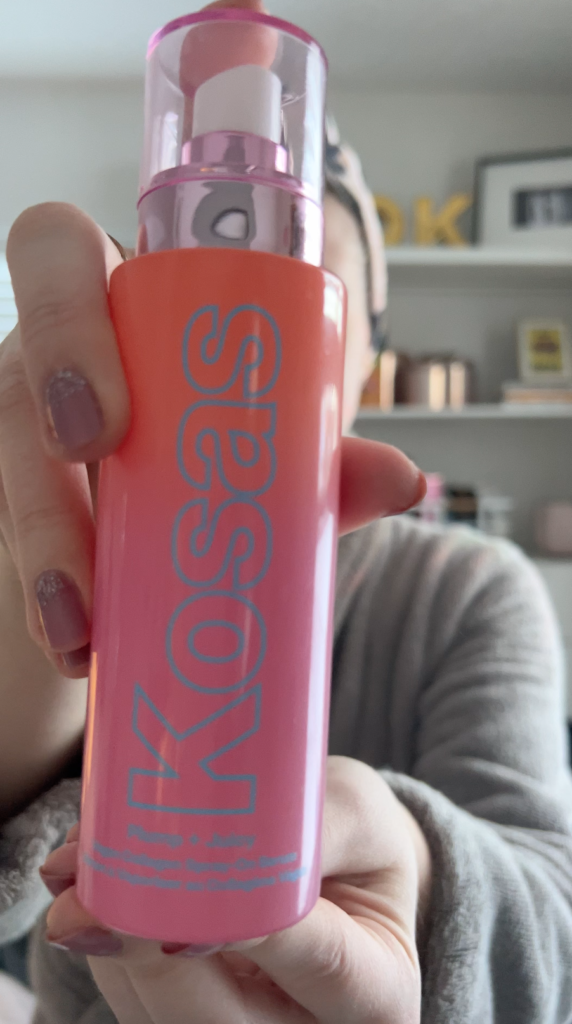 A woman holds up a bottle of Kosas Plump + Juicy Spray.