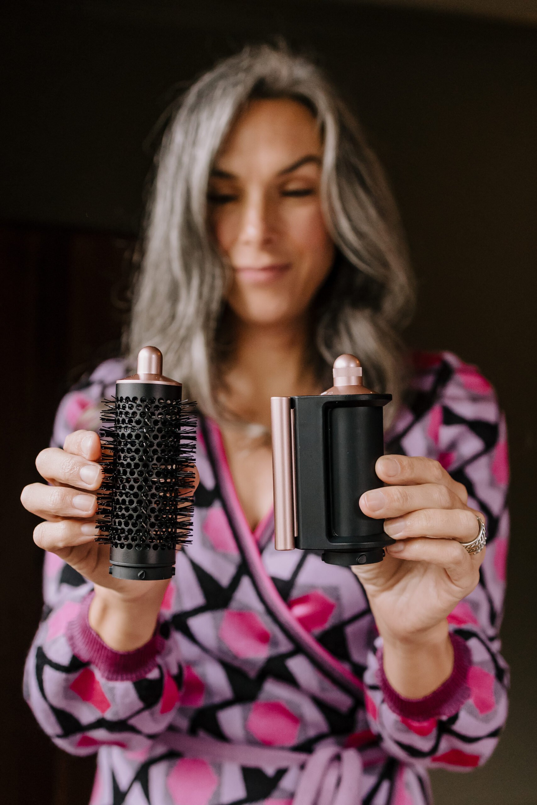 A woman holds up two Dyson Airwrap attachments: The round volumizing brush (left) and the Coanada smoothing dryer (right).
