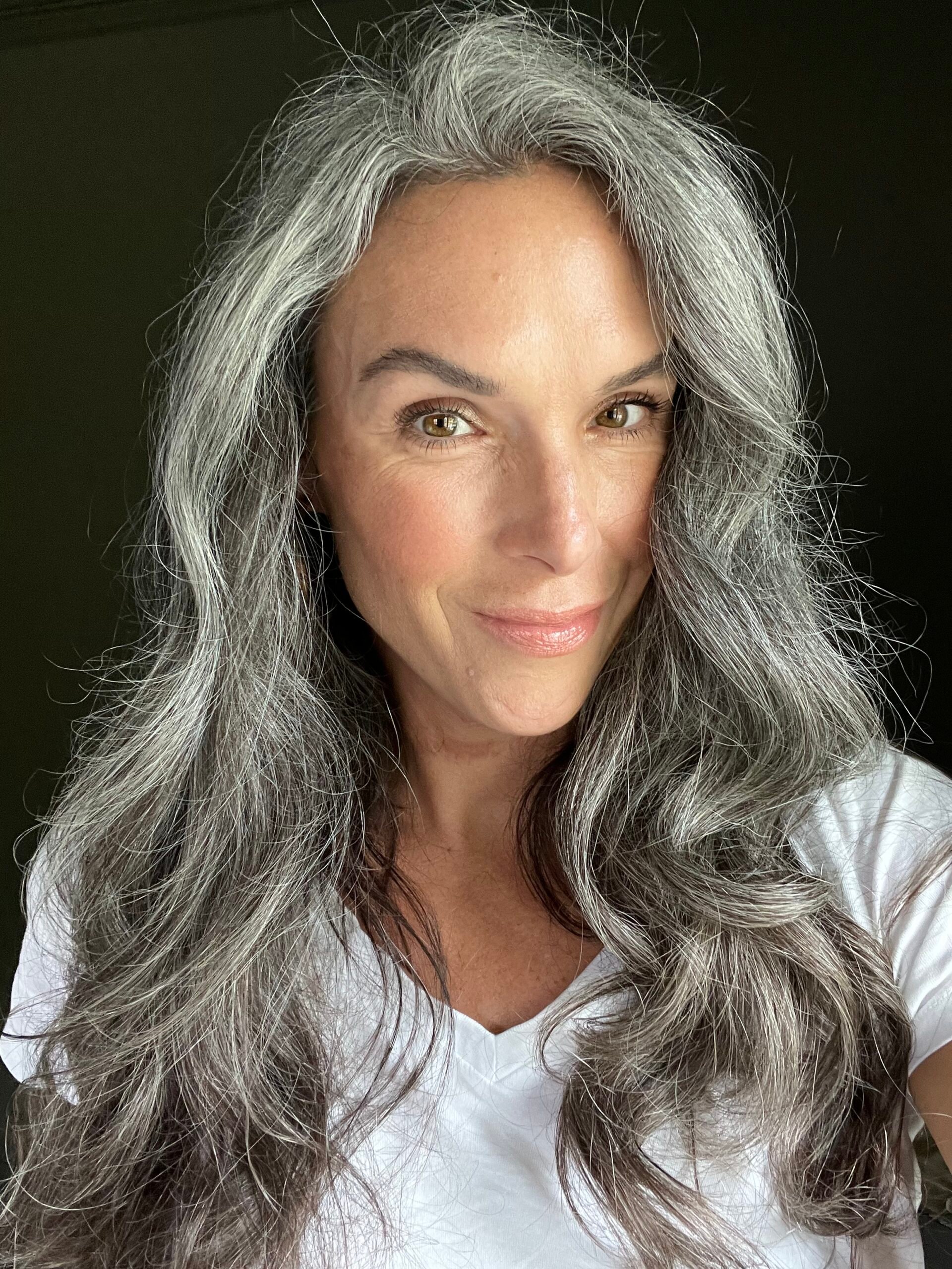 A woman with long wavy gray hair wears RMS blush. 