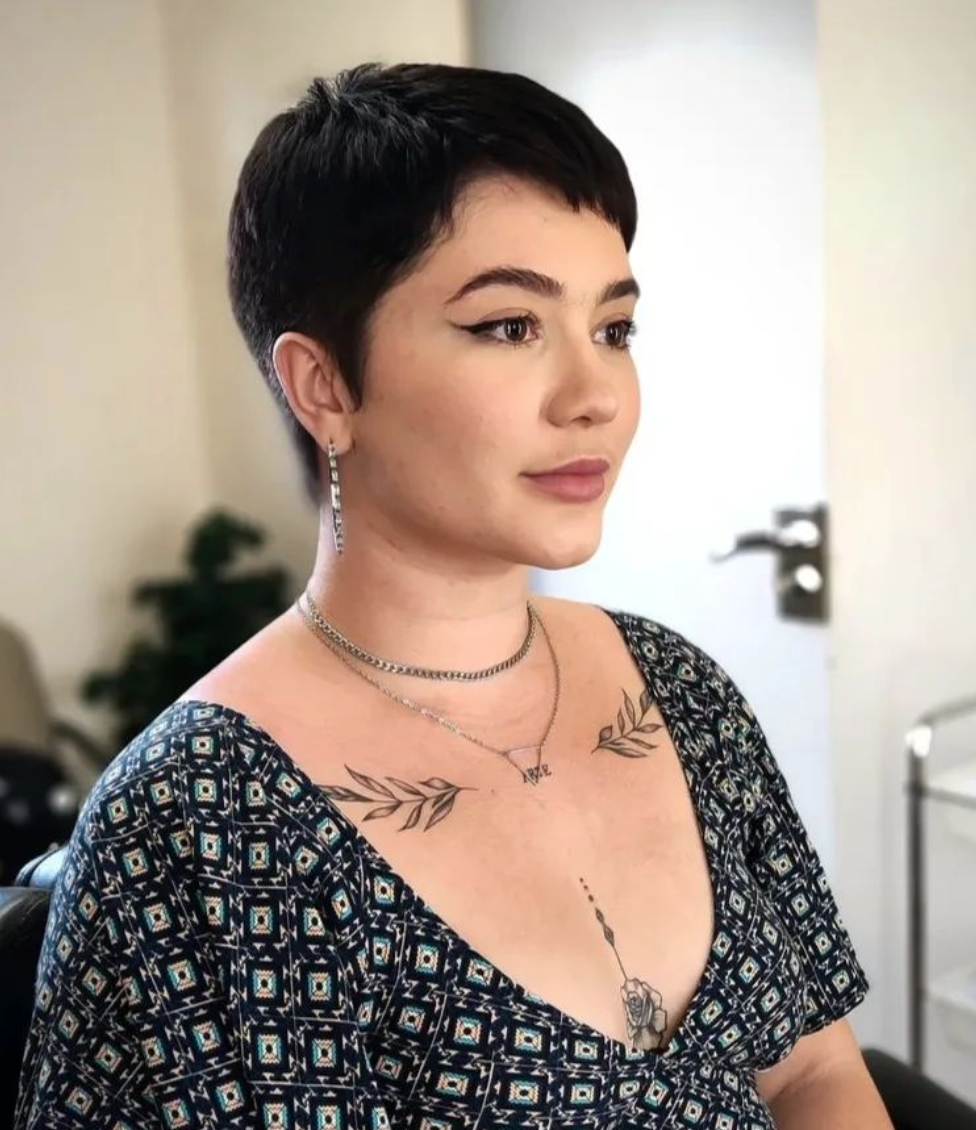 A woman with a black pixie cut and botanical tattoos on her chest looks off to the side. 