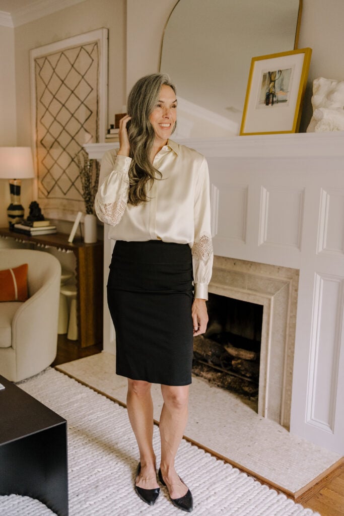 A woman standing in a living room while wearing a black pencil skirt and a white silk top.