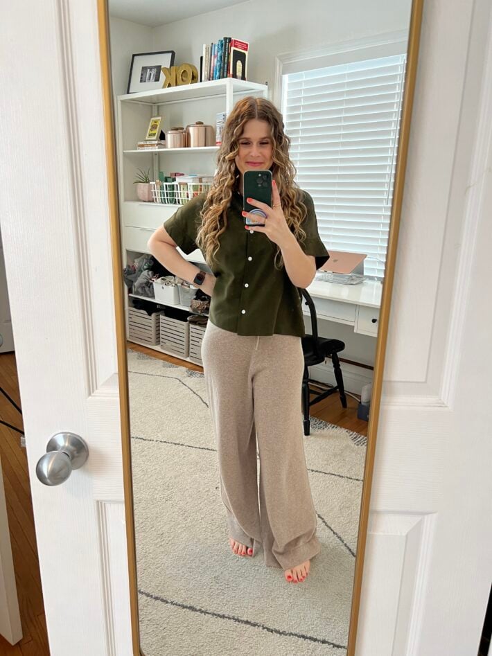 A woman standing in an office wearing an olive silk shirt and cashmere pants. 