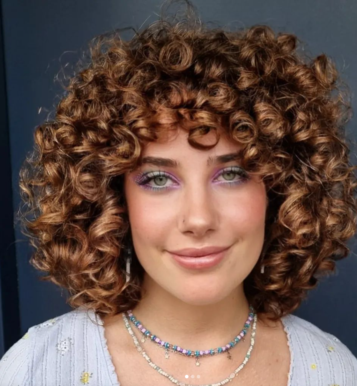 A woman with curly brown hair and purple eyeshadow smiles. 