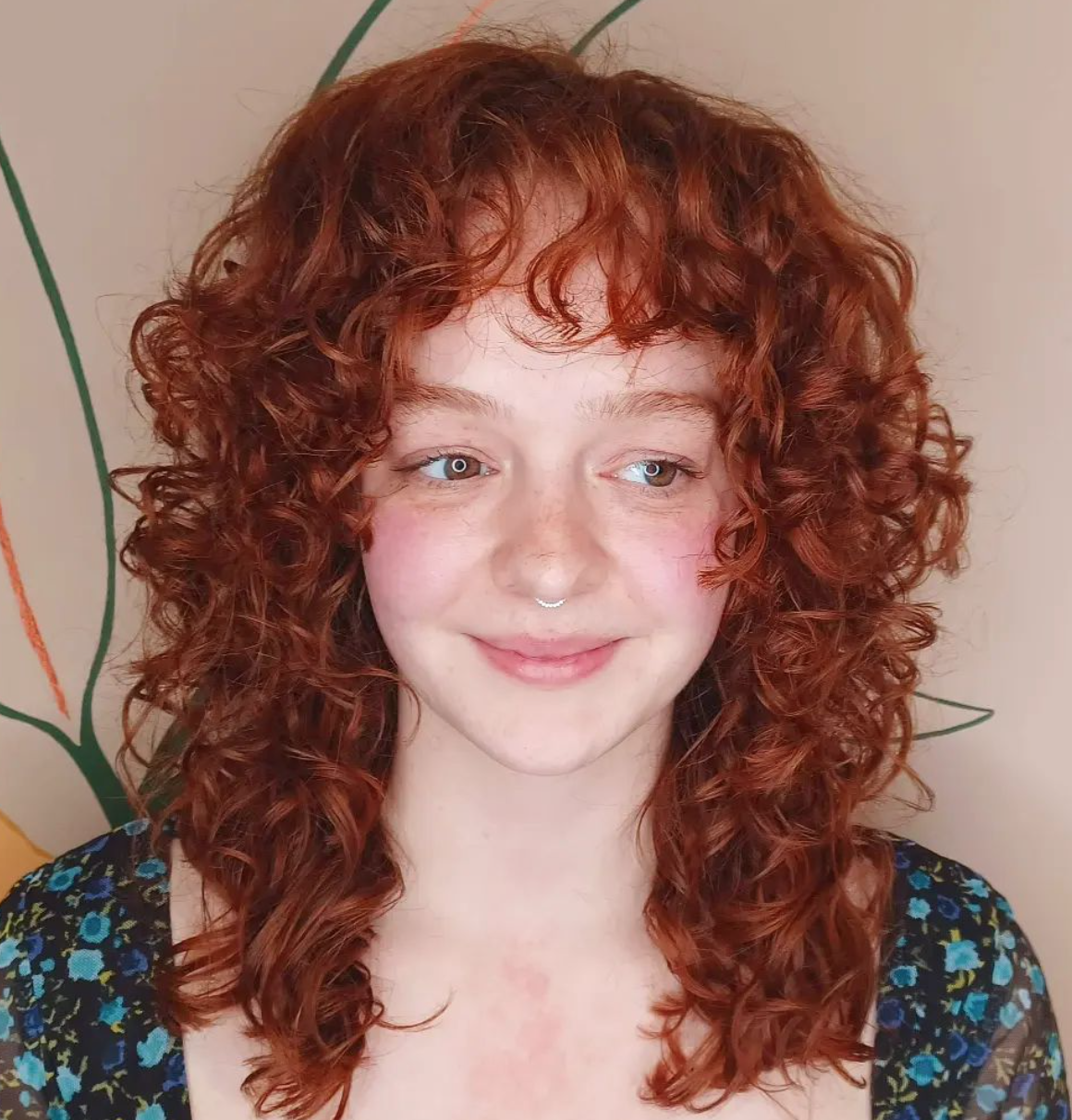 A smiling woman with curly red hair and a nose ring rocks a wolf cut.