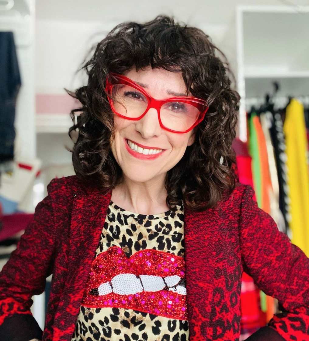 A smiling woman with bright red glasses looks onward.
