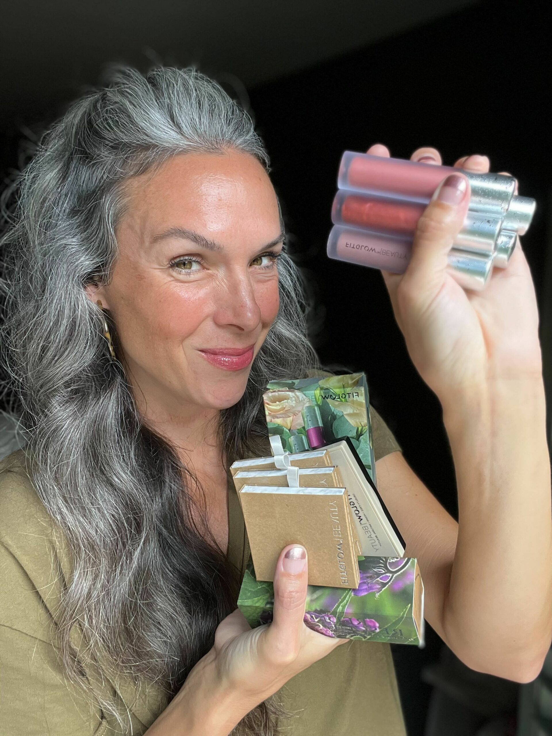 A woman with curly gray hair holds up products from Fitglow Beauty.