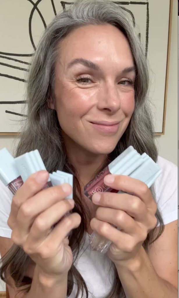 A woman smiling while holding up Kosas Wet Lip Oil Gloss.
