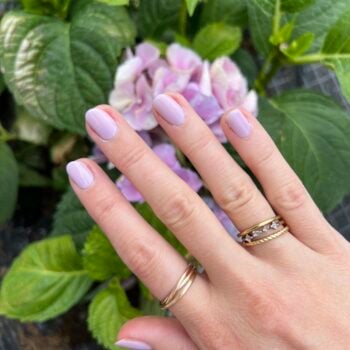A woman's hand with gold rings and lavender nail polish is stretched out over purple flowers.