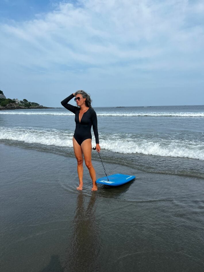 A woman on the beach with a boogie board in a black swimsuit with long sleeves. 