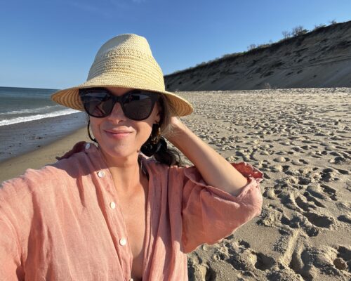 A woman stands on a beach with a hat on her head.