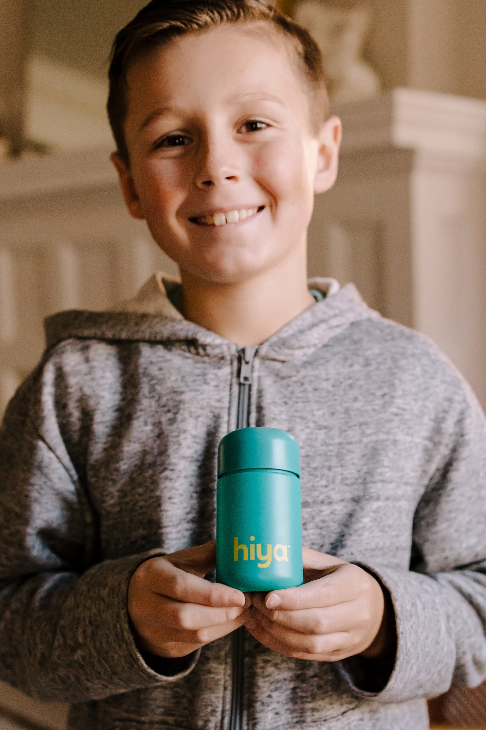 A smiling boy holding up Hiya probiotics in a teal bottle. 